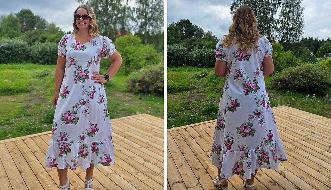 Kvinna står utomhus på en altan med grönt landskap i bakgrunden, iklädd en blommig A-linjeformad Lottaklänning med puffärm och volangkant. Bilden visar både fram- och baksidan av klänningen.